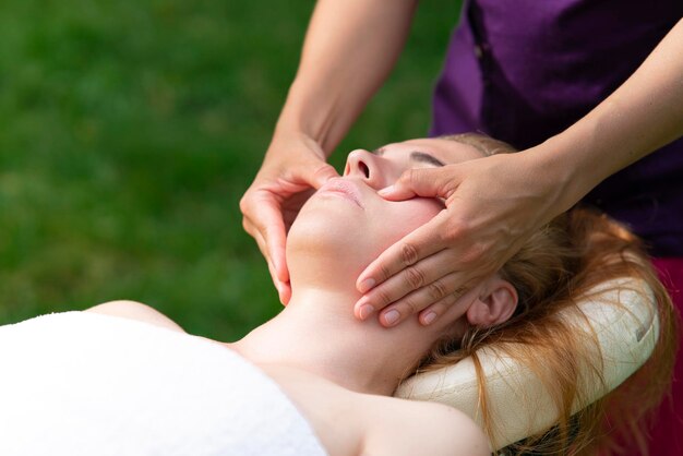 Kopf der jungen Frau, der Gesichtsmassage mit Doktorhänden im Freien auf natürlichem Hintergrund erhält, genießt, sich entspannt. Hübsche weibliche entspannte Patientin, die Schönheitsbehandlung, gesunde Haut, Anti-Aging-Verfahren macht.