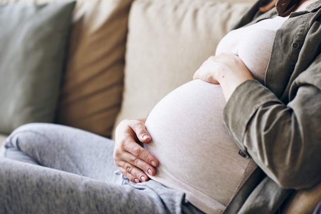 Kopfloser Schuss der erholsamen schwangeren Frau mit ihren Händen auf beiden Seiten des Bauches entspannend auf der Couch mit Kissen an der Wand