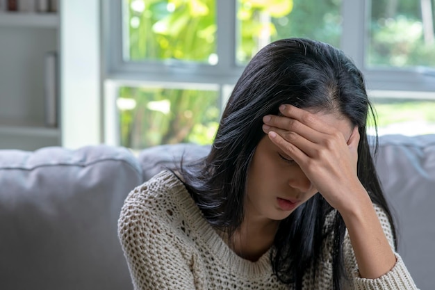 Kopfschmerzen Junge asiatische Frau sitzt mit geschlossenen Augen auf einem Sofa und berührt ihren Kopf, während sie an einer Migräne leidet