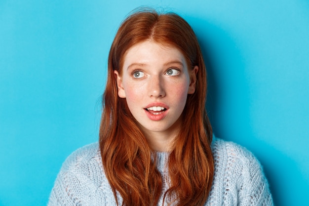 Kopfschuss des nachdenklichen rothaarigen jugendlich Mädchens, das die obere linke Ecke betrachtet und auf das Logo mit neugierigem Ausdruck starrt und über blauem Hintergrund steht.