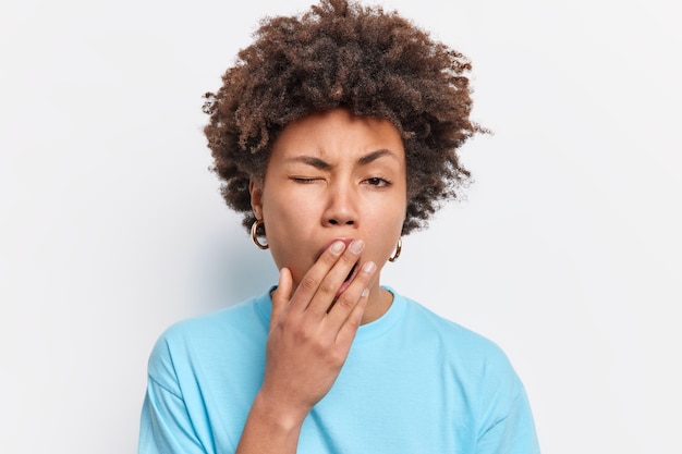 Kopfschuss einer jungen afroamerikanischen schläfrigen Frau, die gähnt, hat einen erschöpften Ausdruck, der müde ist von allem, was die Arbeit satt hat, trägt ein lässiges blaues T-Shirt, das über weißer Wand isoliert ist. Energiemangel