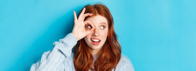 Kopfschuss eines hübschen rothaarigen Mädchens im Pullover, das nach links auf die Promo blickt, mit einem Okay-Schild über dem Auge, das ag steht
