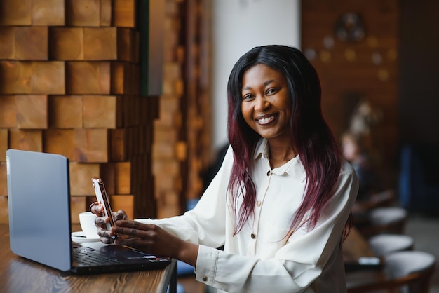 Kopfschussporträt einer glücklich lächelnden afroamerikanischen Frau, die am Tisch im Café sitzt und in die Kamera schaut, aufgeregte Frau, die am Computer arbeitet und Hausaufgaben macht, um einen Bericht im Kaffeehaus vorzubereiten