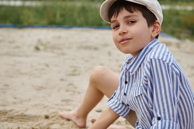 Kopfschussporträt eines entzückenden Schuljungen, der beim Spielen auf dem Sand während der Sommerferien in die Kamera schaut. Sommerferien Konzepte. Sommer Camp