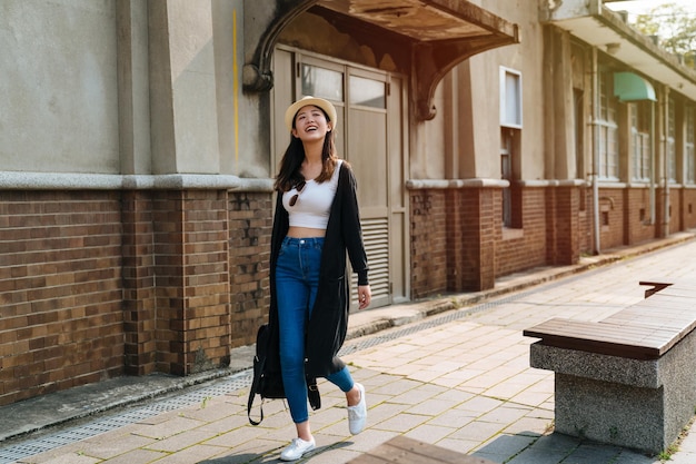 kopieren sie raum in voller länge junge asiatische koreanerin tragen strohhut und tragetasche zu fuß in der alten stadt. fröhliche dame, die zur seite lächelt, während sie sich bei sonnenuntergang im freien entspannen. glückliche Frau in bauchfreiem Oberteil und Jeans