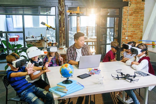 Koreanischer Lehrer mit sechs kaukasischen Schülern, die Augmented-Reality-VR-Brillen in einem Informatik-Grundschulunterricht verwenden