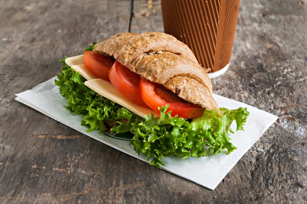 Korn vegetarisches Croissant-Sandwich und eine Tasse Kaffee aus Pappe auf einem alten Holzhintergrund