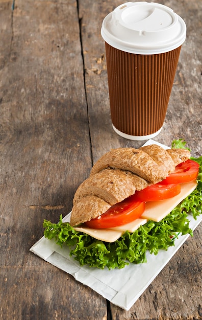 Korn vegetarisches Croissant-Sandwich und eine Tasse Kaffee aus Pappe auf einem alten Holzhintergrund