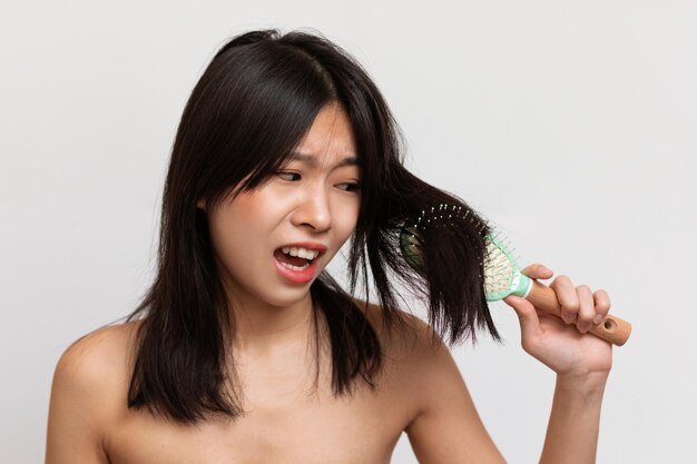 Kosmetik für trockenes und sprödes Haar Gereizte koreanische Dame kann ihr Haar nicht kämmen, das über weißem Studiohintergrund steht