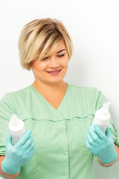 Kosmetikcremes und Hautpflegeprodukte in den Händen der weiblichen Kosmetikerin, die lächelt und über dem weißen Wandhintergrund steht