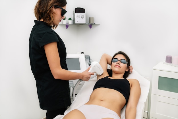 Kosmetikerin, die einer Frau in der Achselhöhle, in der sie eine Laserschutzbrille trägt, eine Laserdiodenbehandlung gibt