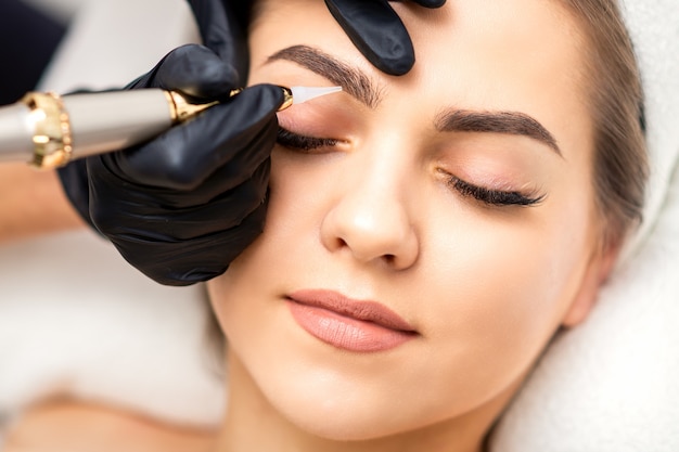 Kosmetikerin, die permanentes Make-up auf Augenbrauen der jungen Frau durch spezielle Tätowiermaschine aufbringt