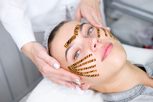 Kosmetikerin, die Taping-Gesichtsverfahren unter Verwendung von tigerfarbenen Bändern im Schönheitssalon macht