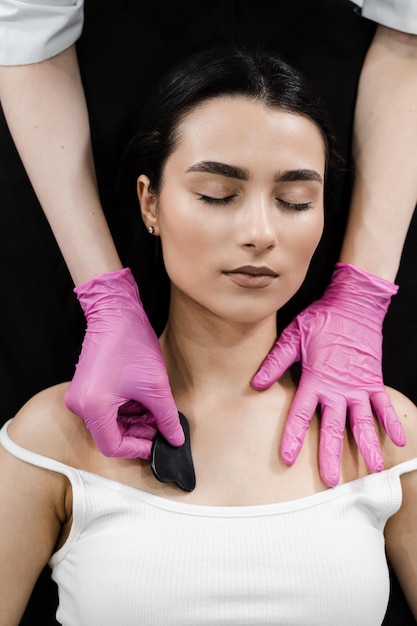 Kosmetikerin führt eine Gua-Sha-Massage durch, um die Durchblutung zu verbessern und die Gesichtshaut zu straffen. Gouache-Massage für das Gesicht eines Mädchens mit einem Steinschaber