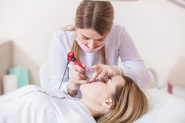 Kosmetikerin macht permanentes Make-up der Lippen einer jungen Patientin im Büro