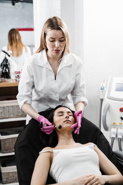 Kosmetikerin verwendet Steinroller für die Gesichtsmassage einer Patientin. Gesichtsmassage mit Kosmetikroller in Kosmetikklinik. Prozess der Gesichtsmassage mit schwarzem Steinroller in Nahaufnahme