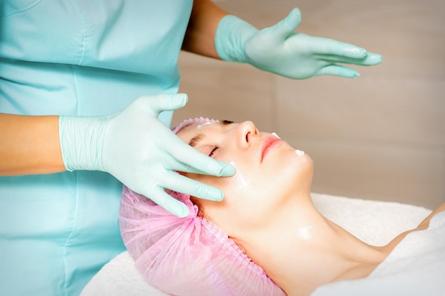 Kosmetologe mit behandschuhten Händen trägt eine feuchtigkeitsspendende Maske mit Peeling-Creme auf das weibliche Gesicht auf Kosmetische Gesichtsbehandlung Verfahren zur Gesichtspflege