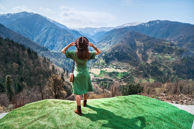 Kostenlose glückliche Boho-Chic-Reisende mit Hut und grünem langem Kleid, die einen schönen Bergblick und einen guten Lebensmoment genießt