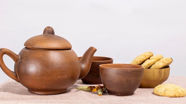 Kräutertee in einer keramischen braunen Teekanne mit Tassen und einer Holzschale mit Keksen auf weißem Tischhintergrund