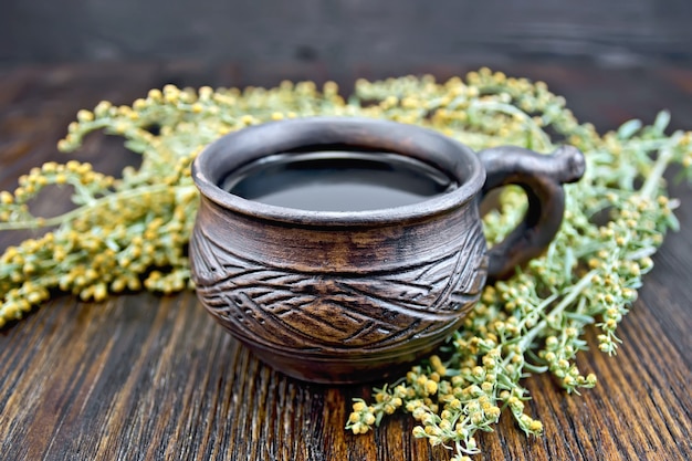 Kräutertee in einer Tontasse, Zweige grauer Beifuß, Wermut getrocknet auf einem Papier auf dem Hintergrund von Holzbrettern