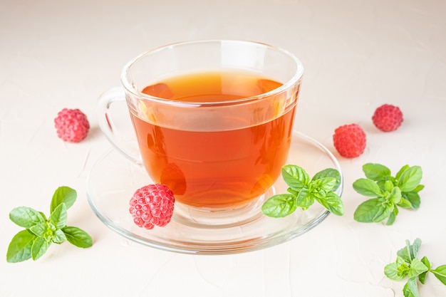 Kräutertee in einer transparenten Tasse mit einer Untertasse auf einem hellen Hintergrund. Rundherum - frische Himbeeren und Minzblätter. Vitamin-Getränke-Konzept.