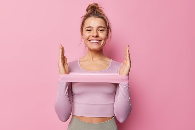 Kraft- und Motivationskonzept. Positive junge Europäerin mit Haarknoten macht Übungen für die Hände und verwendet ein Widerstandsband in Sportkleidung, das glücklich isoliert über rosa Hintergrund lächelt.