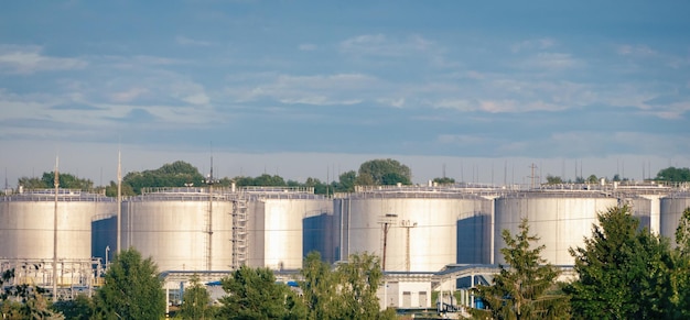 Kraftstofflagertanks sehen die Ölraffinerieanlage im morgendlichen Tageslicht gegen grüne Sommerbäume und blauen Himmel