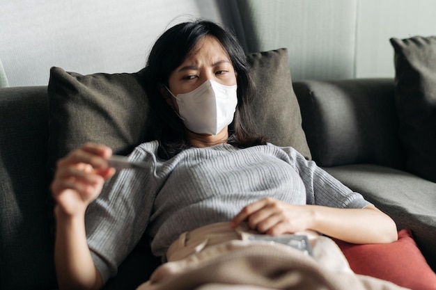 Foto kranke asiatische frau, die klinisches thermometer liest, während sie zu hause auf einem schlafsofa hustet