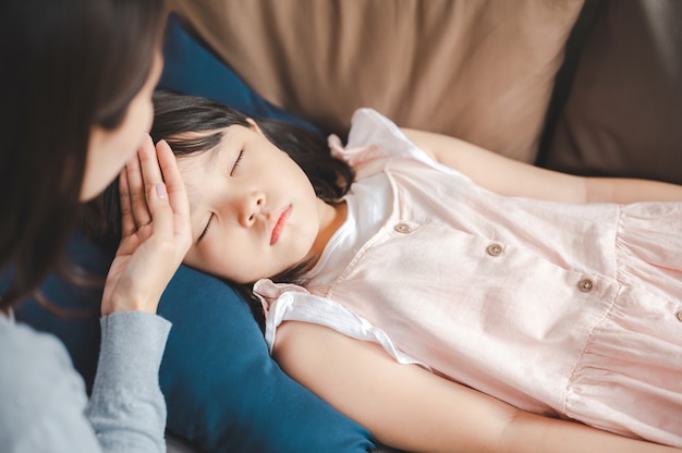 Kranke asiatische Tochter, die mit Fieber auf der Couch schläft, während die Mutter die Temperatur auf ihrer Stirn überprüft