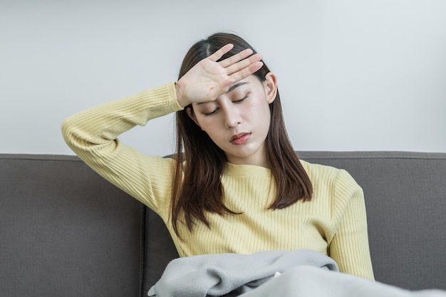 Kranke, Coronavirus covid-19 asiatische junge Frau, Mädchenkopfschmerzen unter der Decke haben Fieber, Grippe und verwenden Taschentücher, die Nase niesen, sitzen auf dem Schlafsofa zu Hause. Gesundheitsversorgung für Viruspersonen.