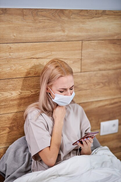 Kranke Frau bekam Fieber gefangen kalt auf dem Bett zu Hause mit Smartphone in den Händen, kranke allergische europäische Frau in der medizinischen Maske mit Allergiesymptomen Husten zu Hause, Grippe-Konzept