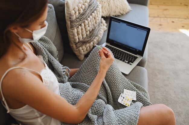 Kranke Frau, die Gesichtsschutzmaske trägt, sitzt auf Couch mit Laptop und hält Thermometer und Pillen während Selbstquarantäne-Selbstisolierung zu Hause.