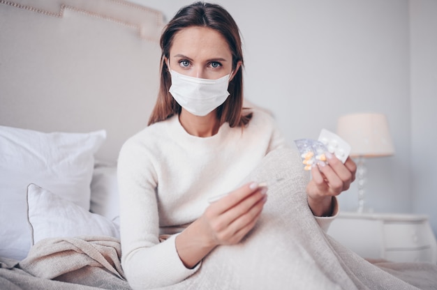Kranke Frau in der Gesichtsschutzmaske, die im Bett liegt und Thermometer und Pillen zu Hause unter Quarantäne hält