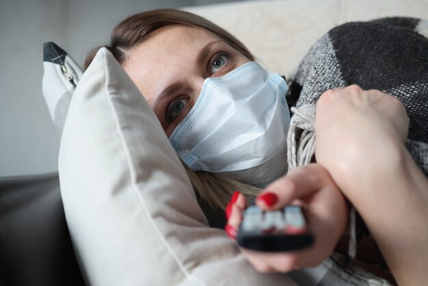 Kranke Frau in medizinischer Schutzmaske liegt auf der Couch und hält Fernbedienung. Coronavirus-Pandemie-Quarantäne-Konzept