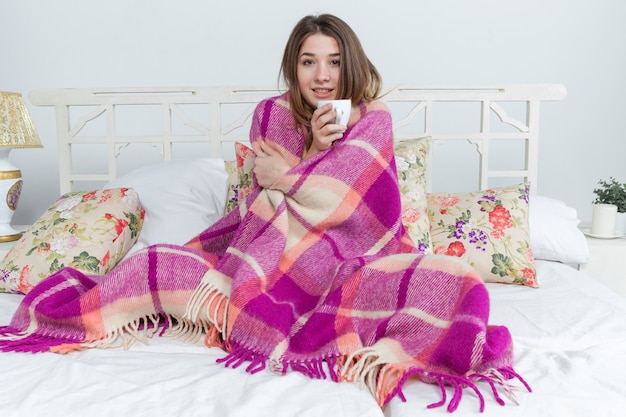 Kranke Frau mit einer Decke bedeckt, die eine Tasse Tee hält, die zu Hause auf der Sofacouch sitzt