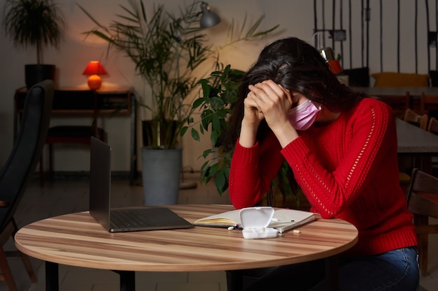 kranke Frau mit Pillen und Thermometer auf einem Tisch, der an einem Laptop von zu Hause auf Quarantäneisolation arbeitet.