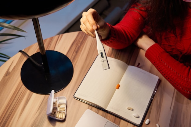 Kranke Frau mit Pillen und Thermometer auf einem Tisch, der an einem Laptop von zu Hause auf Quarantäneisolation arbeitet