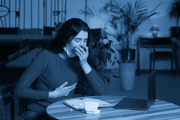 Kranke Frau mit Pillen und Thermometer auf einem Tisch, die von zu Hause aus in Quarantäne an einem Laptop arbeitet