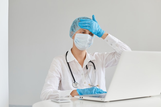 Kranke kranke Ärztin, die ein Kleid, eine chirurgische Maske, eine medizinische Mütze und Handschuhe trägt, in der Klinik vor dem Computer arbeitet und schreckliche Kopfschmerzen und hohe Temperaturen leidet.