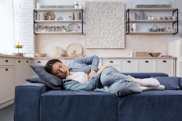 Kranke und traurige frau, die zu hause im wohnzimmer auf dem sofa liegt asiatin hat starke bauchschmerzen