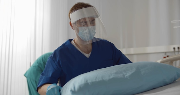 Krankenpfleger in Schutzhandschuhen und Maske arbeitet im Krankenhaus beim Bettenmachen und Wechseln der Bettwäsche