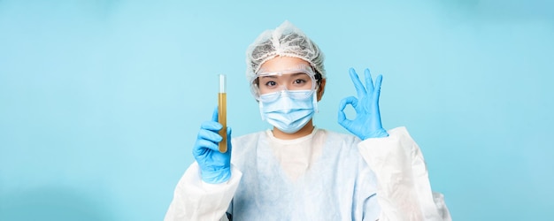 Krankenschwester des Laborpersonals in persönlicher Schutzausrüstung mit Probe im Reagenzglas und Okay-Zeichen t