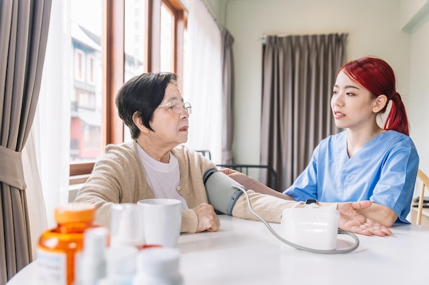 Krankenschwester, die den Blutdruck misst, indem sie ein automatisches Blutdruckmessgerät bei reifen älteren asiatischen Frauen mit Pflege verwendet. Besuch der Pflegekraft zu Hause Konzept der häuslichen Gesundheitspflege und des Pflegeheims