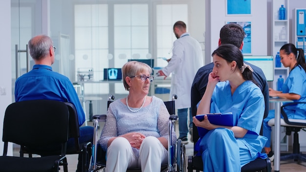 Krankenschwester, die Dokumente einreicht, während sie mit einer behinderten älteren Frau im Wartebereich des Krankenhauses spricht. Patient bittet an der Krankenhausrezeption um Richtung.