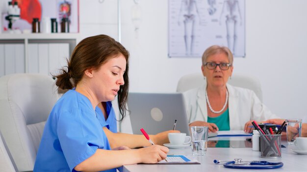 Krankenschwester, die Liste der Patienten während des Brainstormings überprüft, mit Kollegen diskutiert und Notizen in der Zwischenablage macht. Ärzteteam, das im Hintergrund über Krankheitssymptome im Krankenhausbüro spricht.