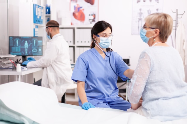 Krankenschwester hört älteres Frauenherz mit Stethoskop im Krankenhaus mit Gesichtsmaske während covid19