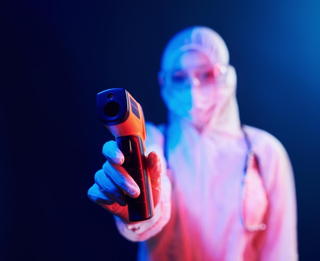 Krankenschwester in Maske und weißer Uniform, die in einem neonbeleuchteten Raum steht und ein Infrarot-Thermometer hält Stoppt die Ausbreitung des Coronavirus