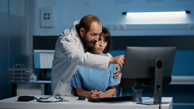 Krankenschwester steht am Schreibtisch und tippt medizinisches Fachwissen in den Computer ein, während der Arzt sie während der Nachtschicht im Krankenhausbüro unangemessen berührt. Assistent, der an einem emotionalen Trauma leidet