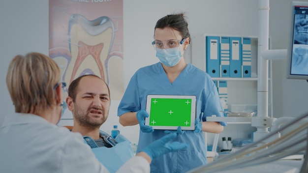 Krankenschwester und Patient analysieren grünen Bildschirm auf einem digitalen Tablet im Zahnmedizinschrank. Zahnmedizinische Fachangestellte, die einen leeren Chroma-Schlüssel mit Mock-up-Vorlage und isoliertem Hintergrund auf einem Kopierraum-Gadget betrachten.