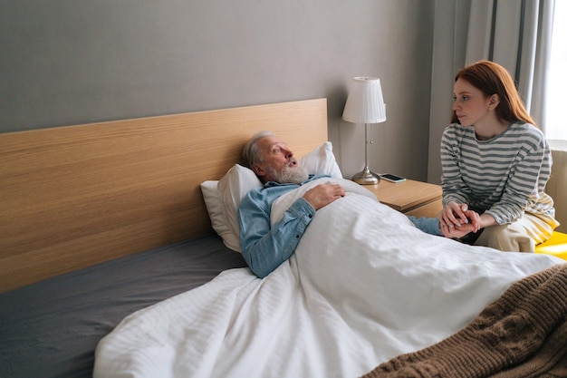 Kranker älterer männlicher Patient, der im Aufwachraum des Krankenhauses auf dem Bett liegt und sich um die Enkelin kümmert. Liebevolle junge Frau, die Hand spricht und tröstenden Krankheitsvater hält